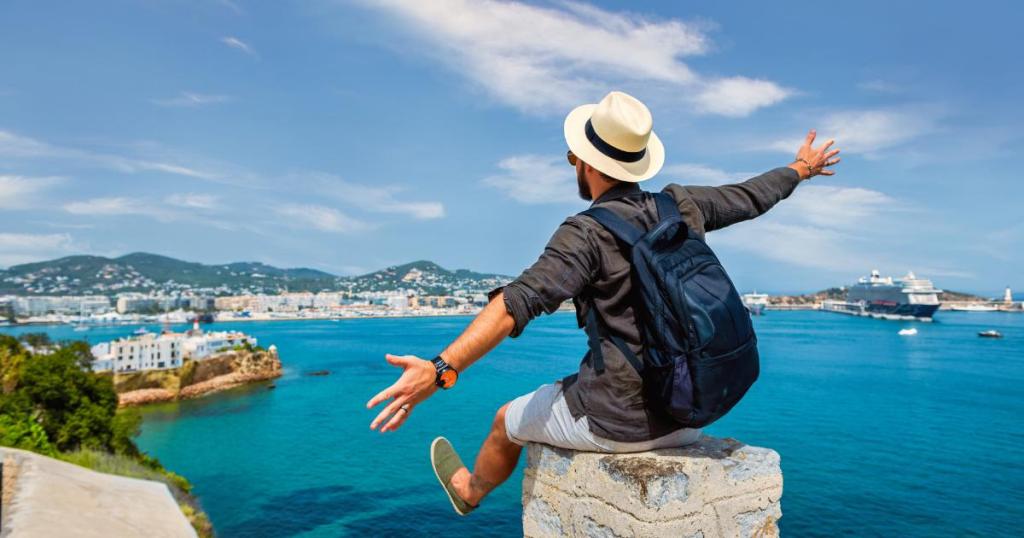 A solo travel man wearing a fedora, sitting on a ledge, gazing at the scenic ocean view with clear blue waters and a calm sky in the background.