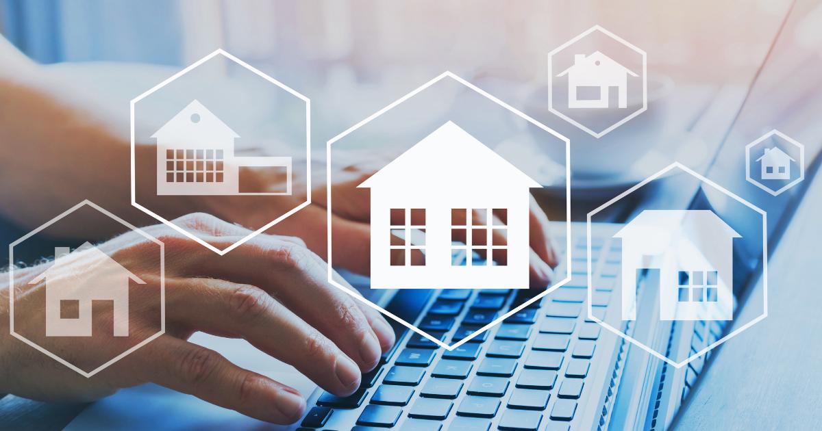 Close-up of hands typing buying a home on a laptop keyboard with graphical illustrations of houses displayed on the screen.